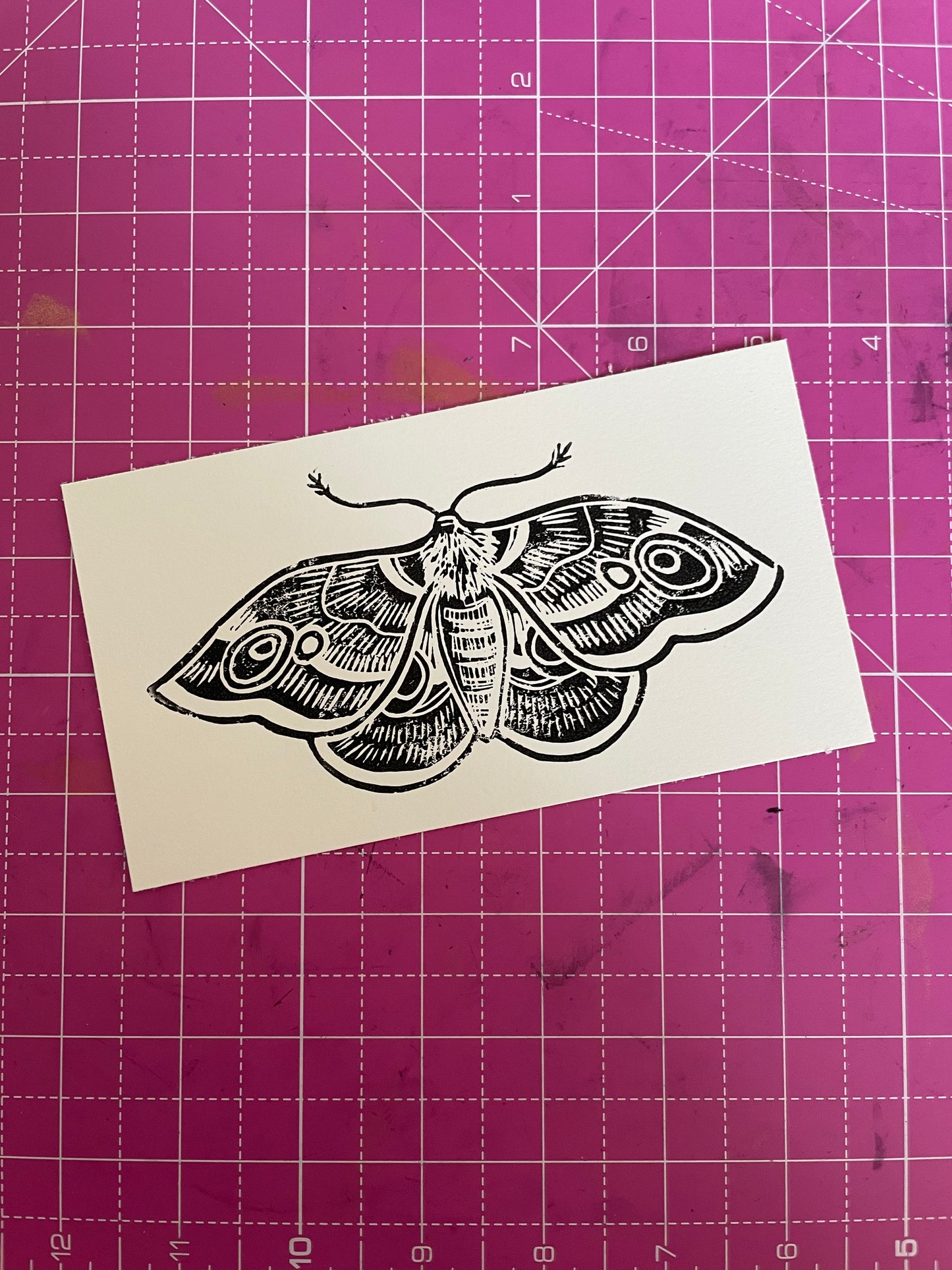 A linocut print of a butterfly moth with black details on a white background, displayed on a cutting mat.