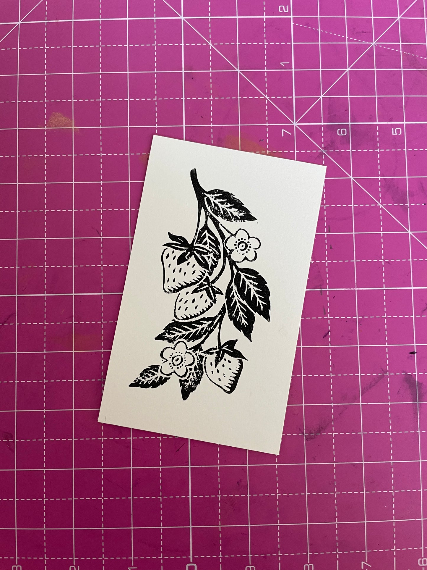 A black and white image of a linocut print featuring a strawberry and leaves on a pink background.
