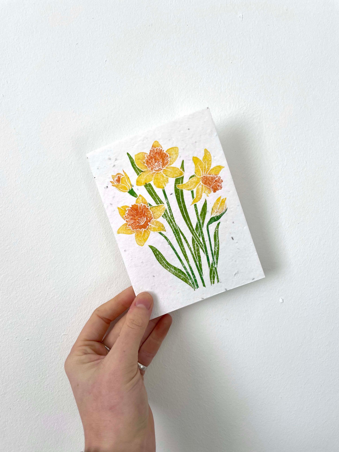 A hand holding a plantable seed card with a lino print design featuring yellow daffodils, accompanied by a recycled Kraft envelope.