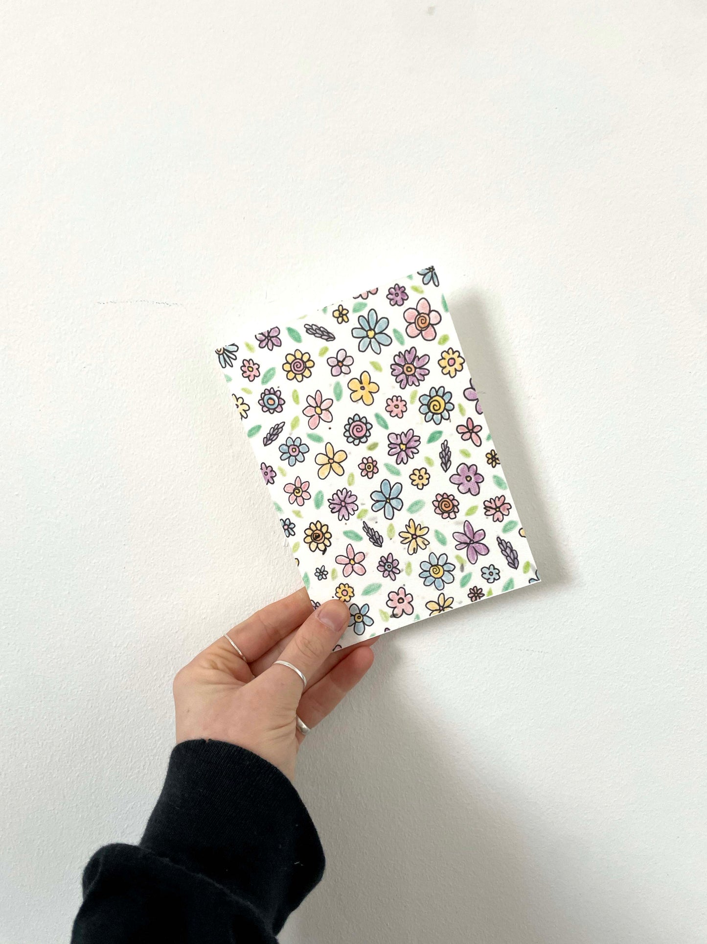 A hand holding a plantable seed card with a floral pattern and a recycled Kraft envelope.